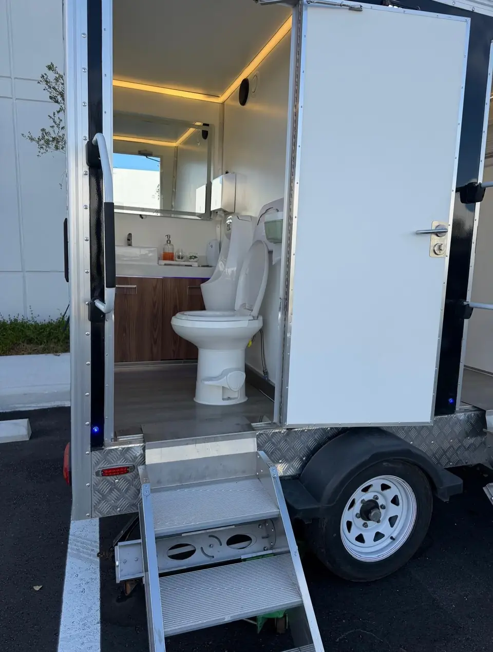 Open restroom trailer door showing a flushing toilet, wood vanity and lit mirror